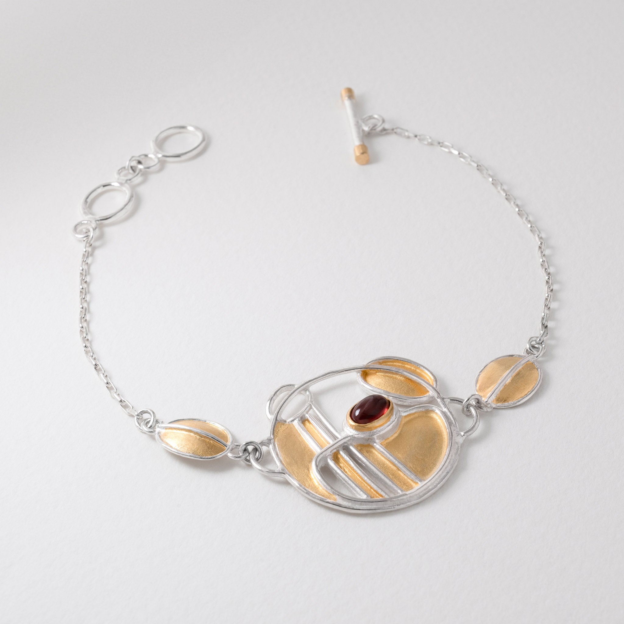 A silver bracelet with gold accents and a central tassel-like charm featuring garnet and gold colors, with a loop at the top for wearing.