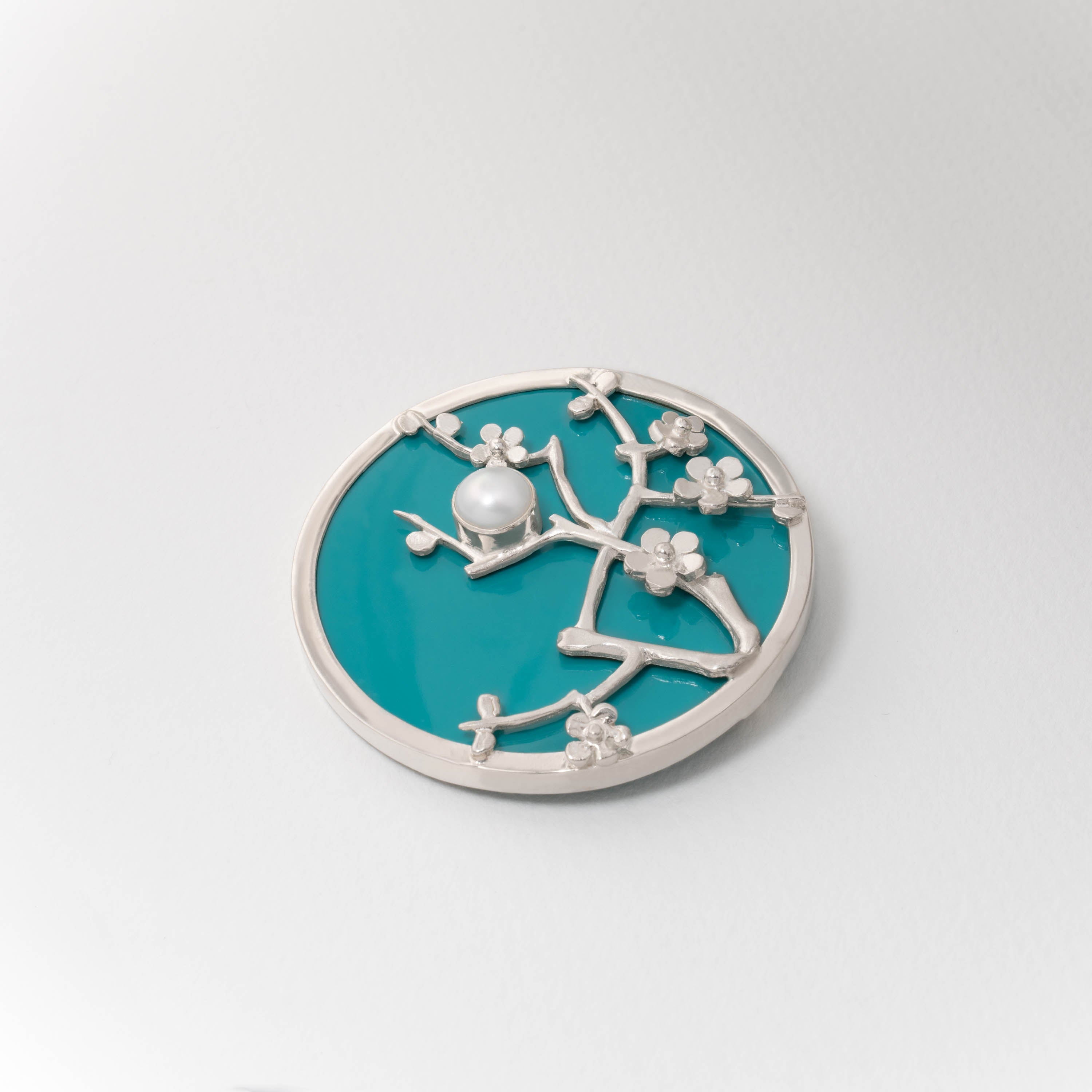 Sterling silver Almond Blossom brooch inspired by Van Gogh. The circular design features silver blossom branches over turquoise blue resin, with a luminous central detail evoking the painter’s impressionist style.